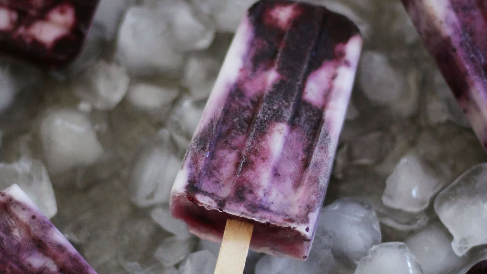 A creamy purple popsicle with a wooden stick rests on ice cubes.