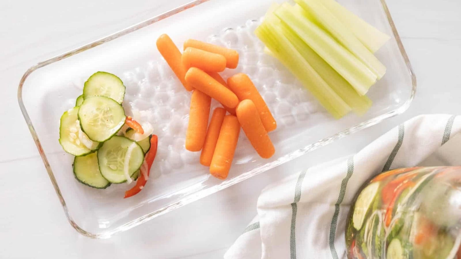 Rectangular glass tray with sliced cucumbers, baby carrots, and celery sticks on a white surface.