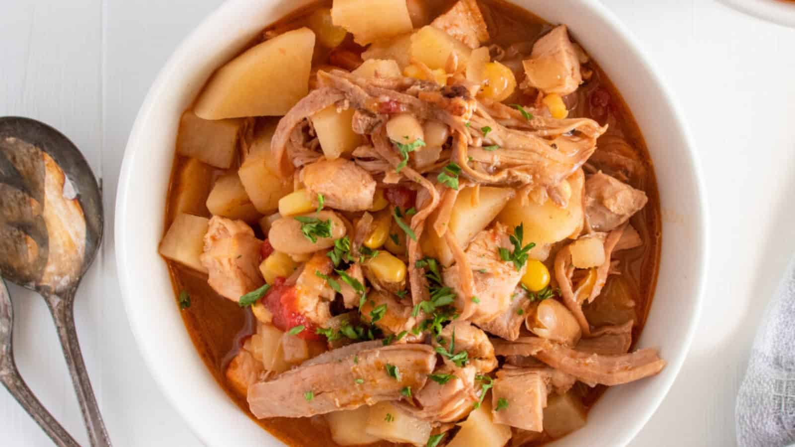 Bowl of chicken stew with potatoes, corn, tomatoes, and herbs, with a spoon on the side.