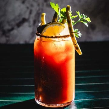 Bloody Mary cocktail in a glass with lemon, green beans, herbs, and a spiced rim on a dark surface.
