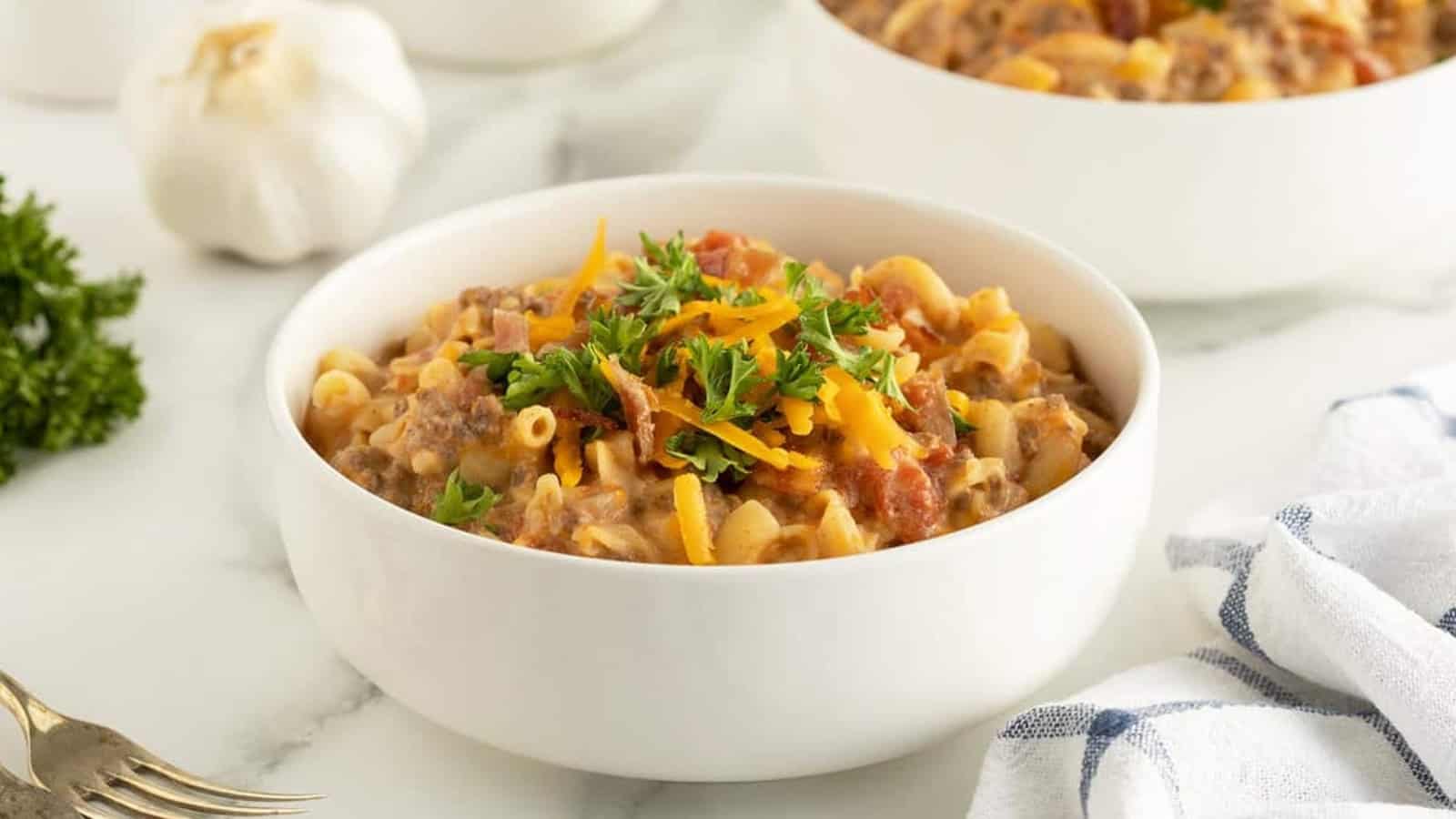 A white bowl of cheesy macaroni with ground beef, topped with parsley and shredded cheese on a marble surface.