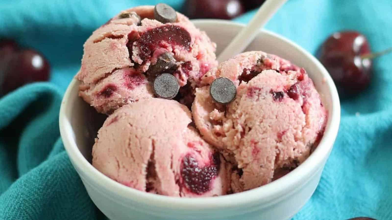 Three scoops of cherry ice cream with chocolate chips in a bowl, with a spoon and cherries in the background.