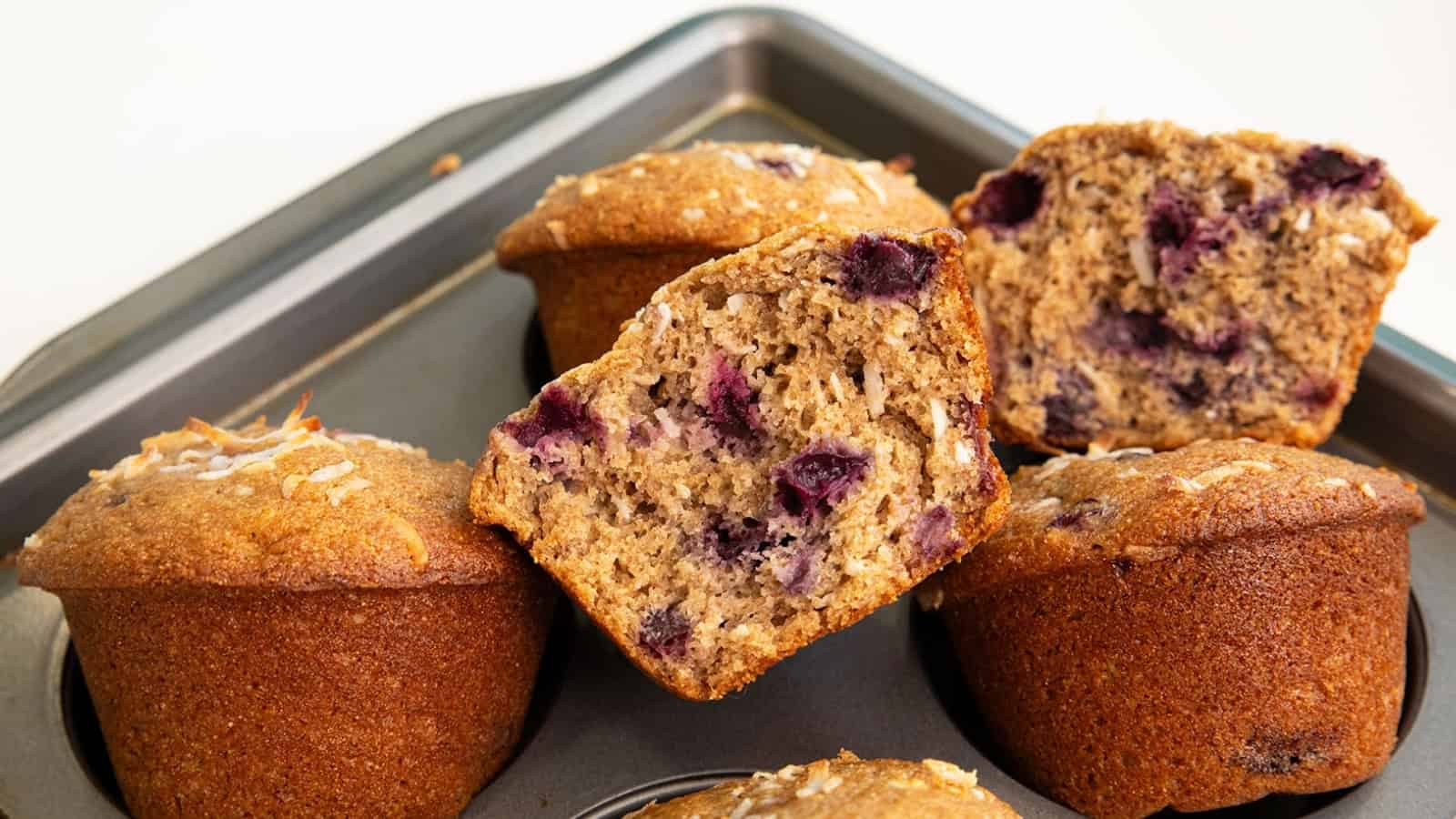 Six blueberry muffins in a muffin tin, with one muffin cut in half showing the inside and berries.