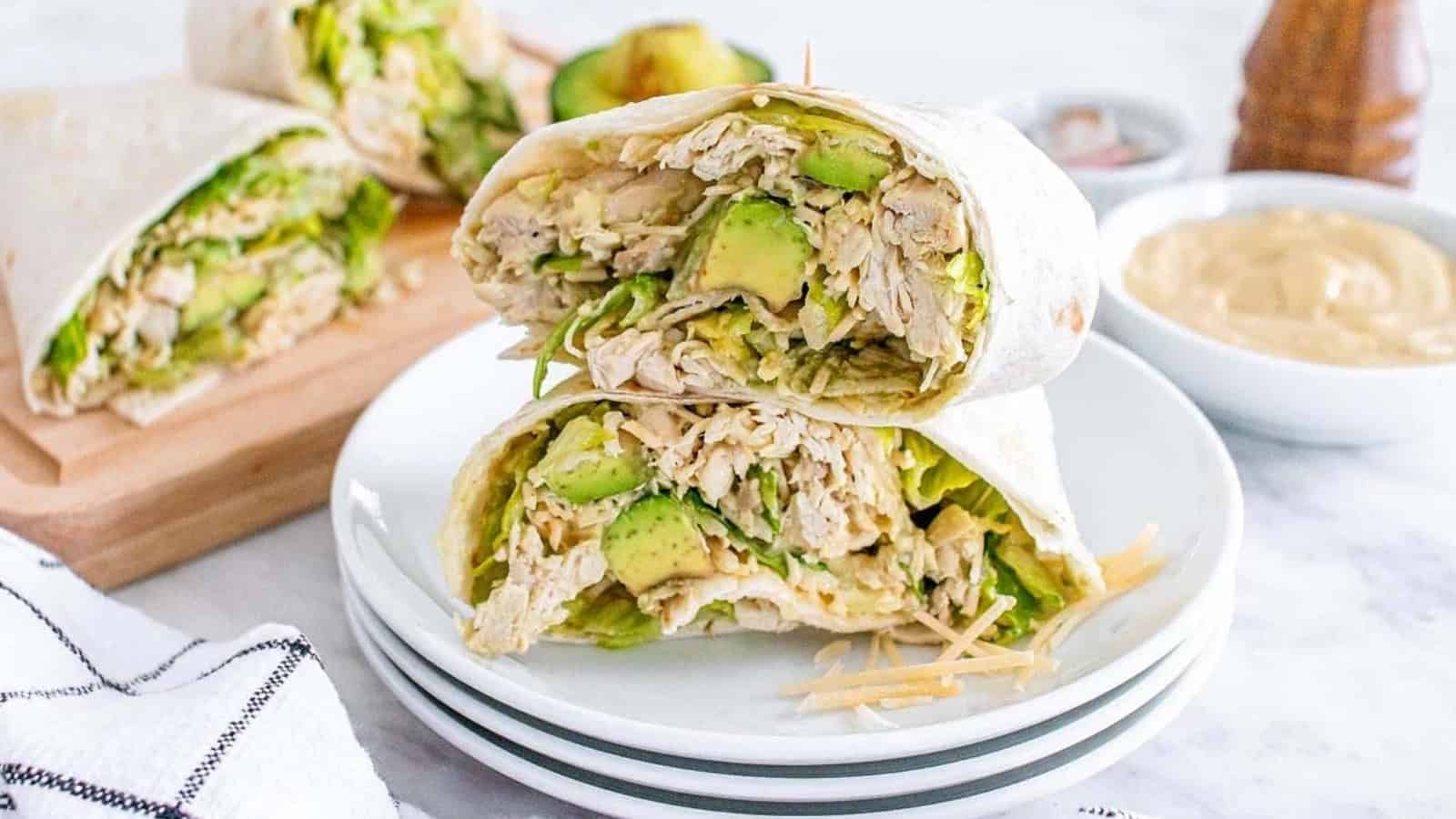 Two chicken and avocado wraps cut in half, stacked on white plates with dip and ingredients in the background.