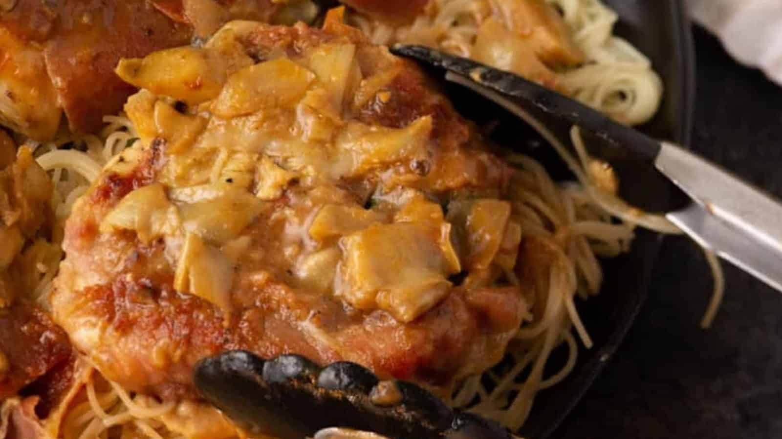 A close-up of pork chops with a creamy artichoke sauce served over a bed of spaghetti noodles.