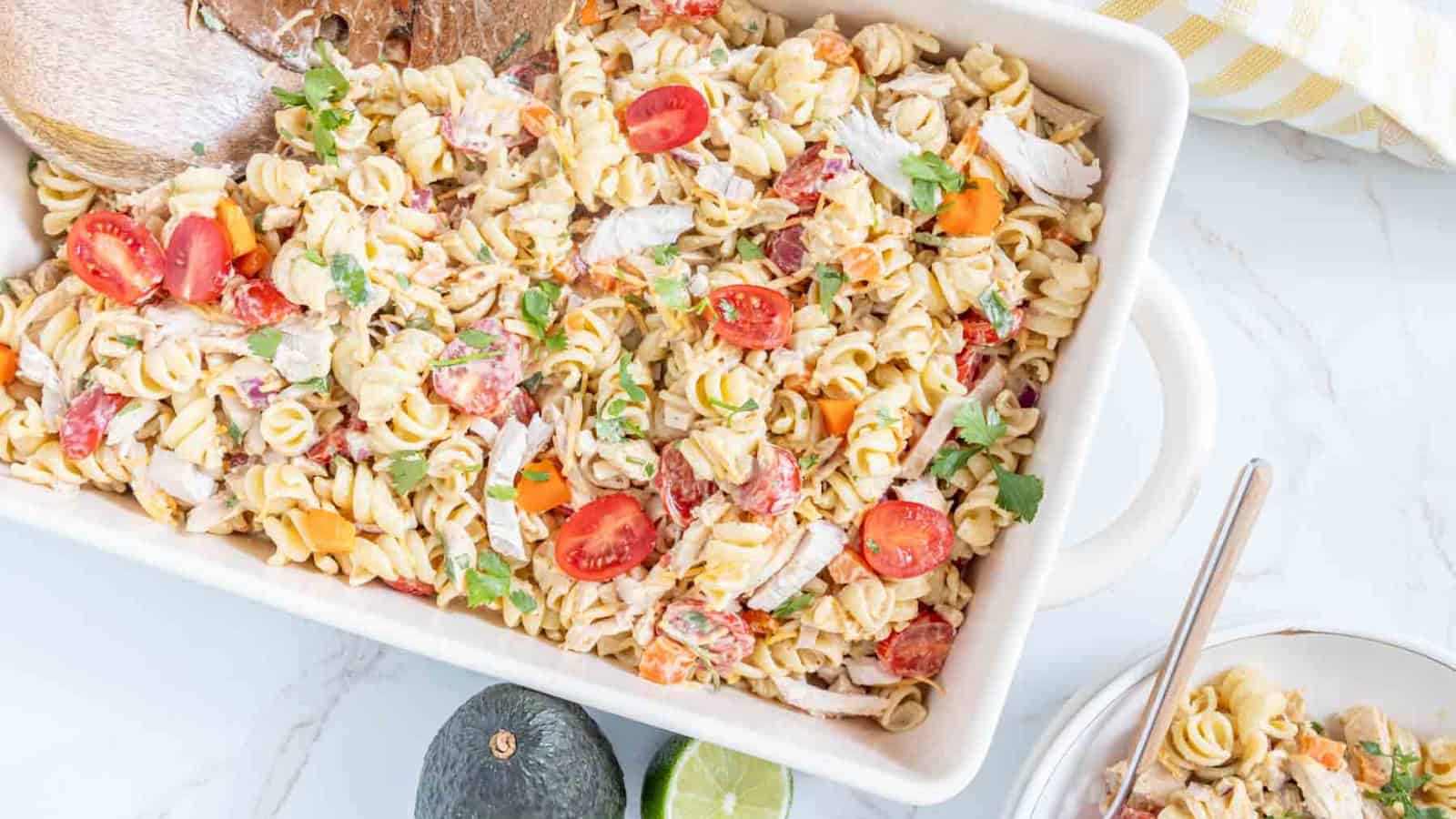 Pasta salad with cherry tomatoes, shredded chicken, and herbs in a white dish, with bread and avocado nearby.