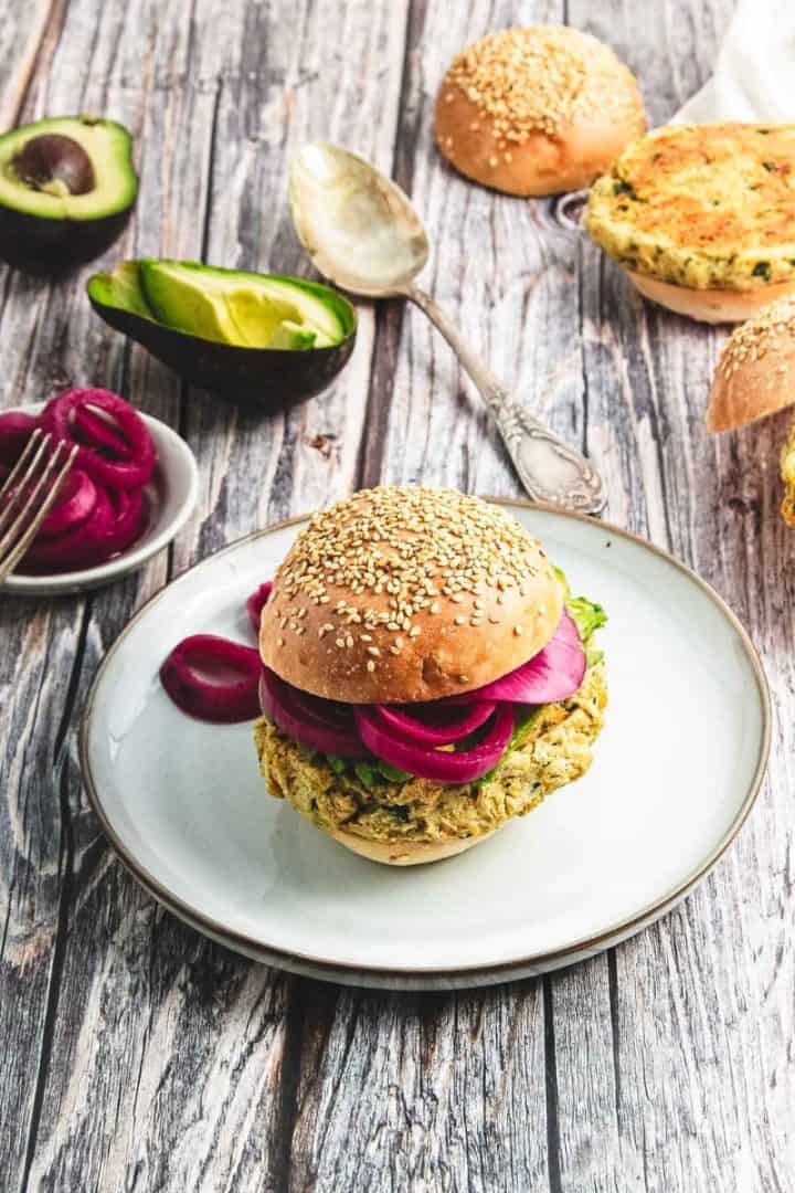 A chickpea burger with pickled onions on a plate, with avocado, buns, and a spoon in the background.