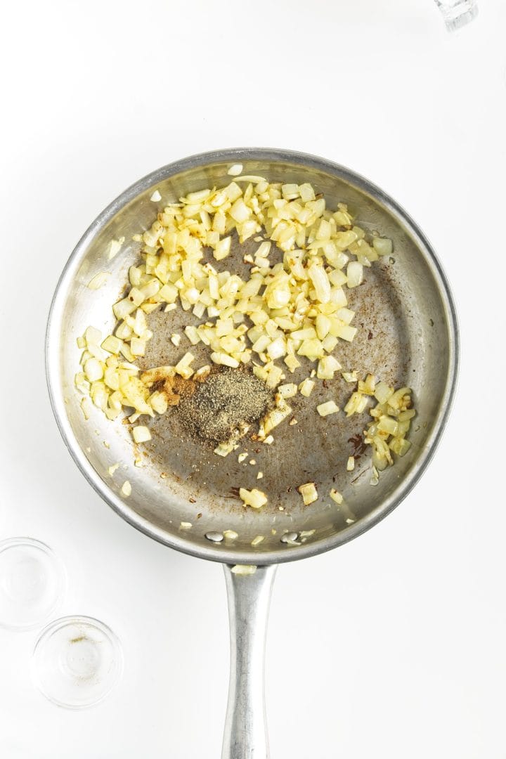 Chopped onions and seasonings cooking in a stainless steel skillet on a white background, perfect for preparing flavorful chickpea burgers.