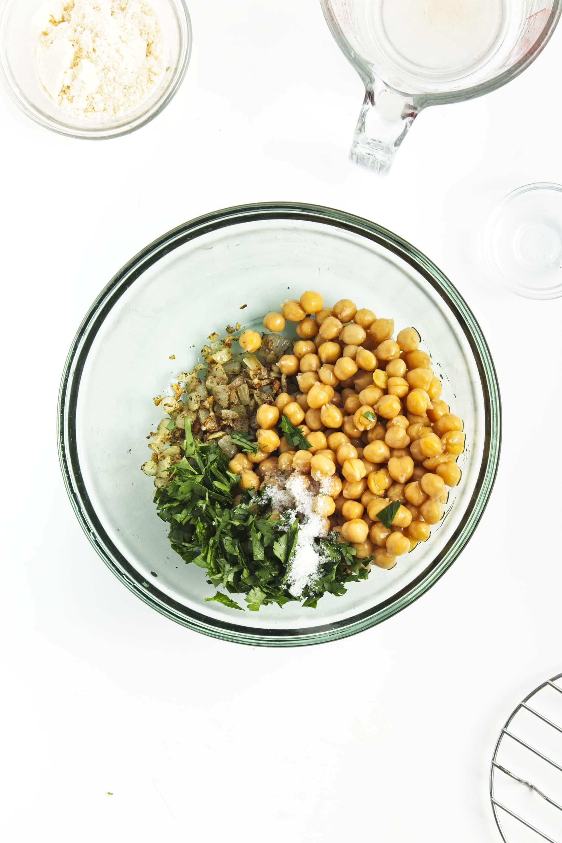 A glass bowl with chickpeas, chopped parsley, salt, and sautéed onions on a white surface-perfect ingredients for homemade chickpea burgers.