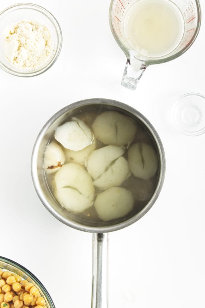 A pot of peeled potatoes boiling in water, surrounded by bowls and a measuring cup on a white surface-perfect for prepping sides or chickpea burgers.