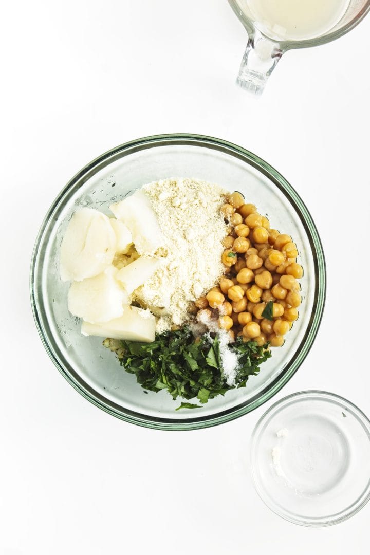 Glass bowl with ingredients for chickpea burgers-chickpeas, flour, potatoes, chopped herbs, and spices-on a white surface.