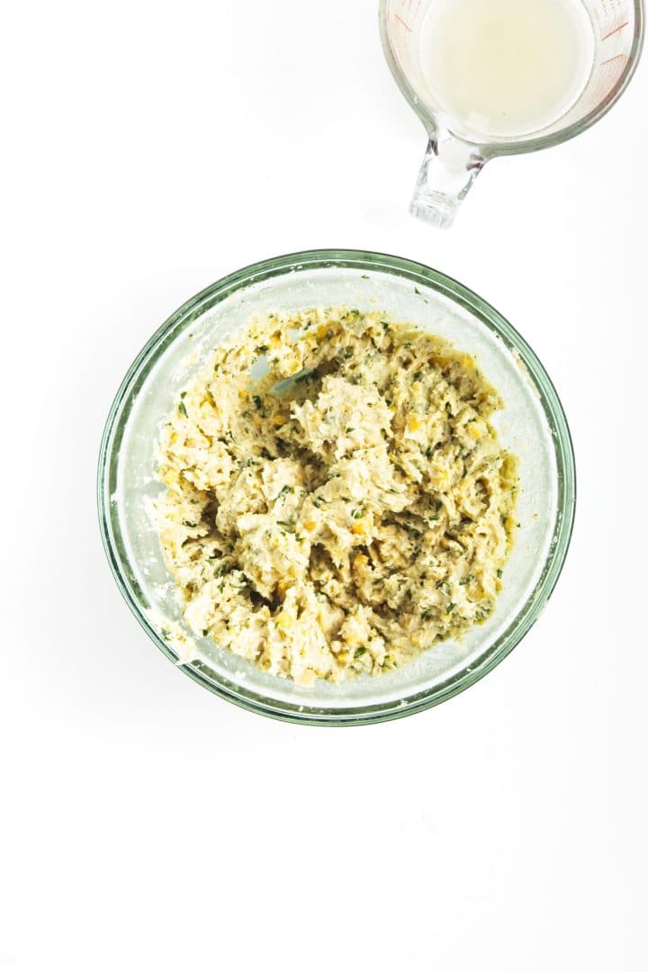 A glass bowl of herbed mixture for chickpea burgers sits next to a measuring cup with liquid on a white background.