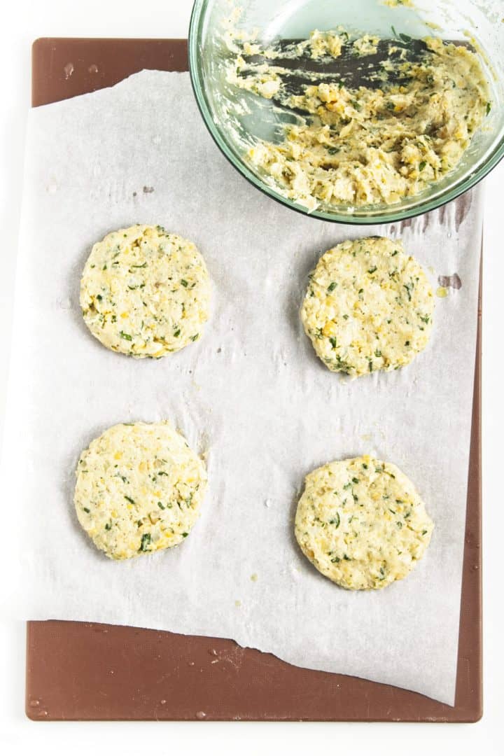 Four uncooked chickpea burgers rest on parchment paper beside a bowl of mixed ingredients on a brown board.