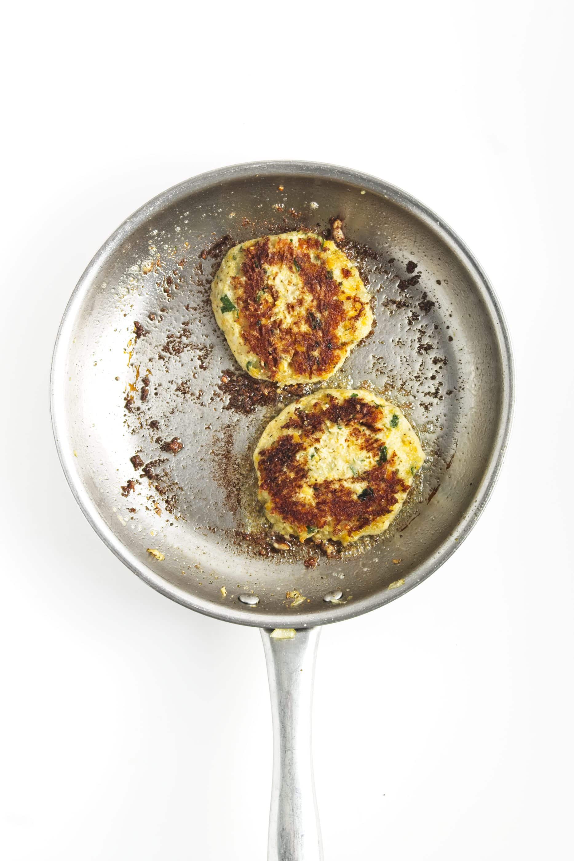 Two browned chickpea burgers sizzle in a stainless steel frying pan on a white background.