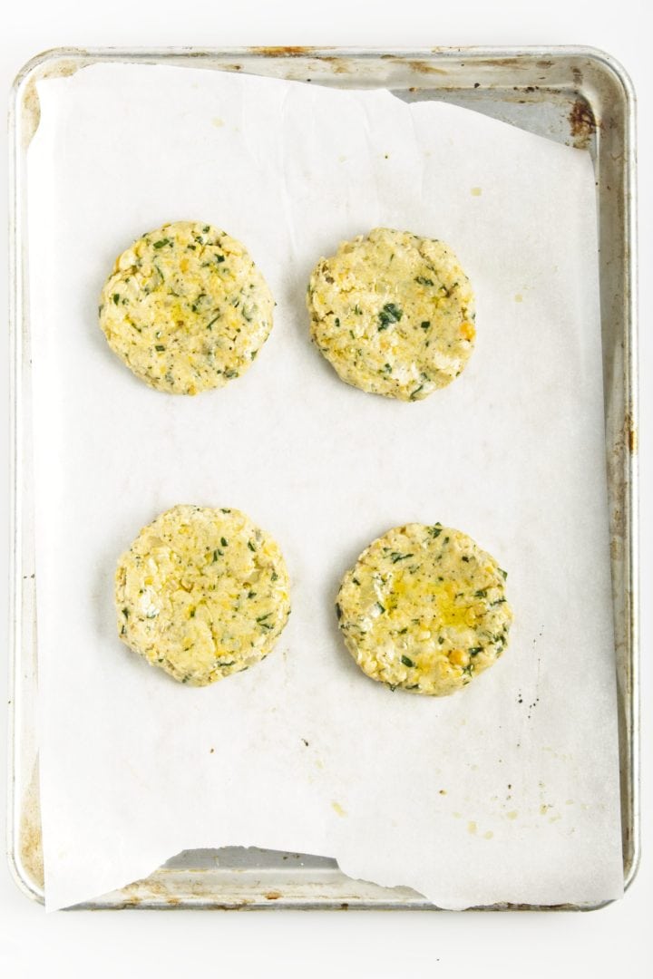 Four uncooked chickpea burgers rest on a parchment-lined baking sheet, viewed from above.