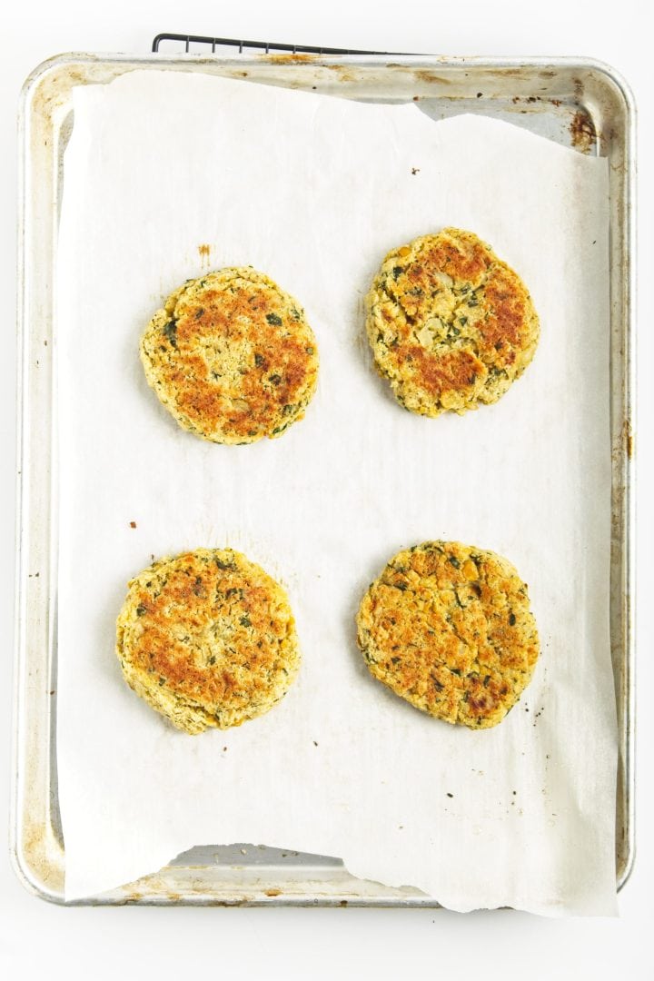 Four baked chickpea burgers rest on parchment paper atop a metal baking sheet, viewed from above.
