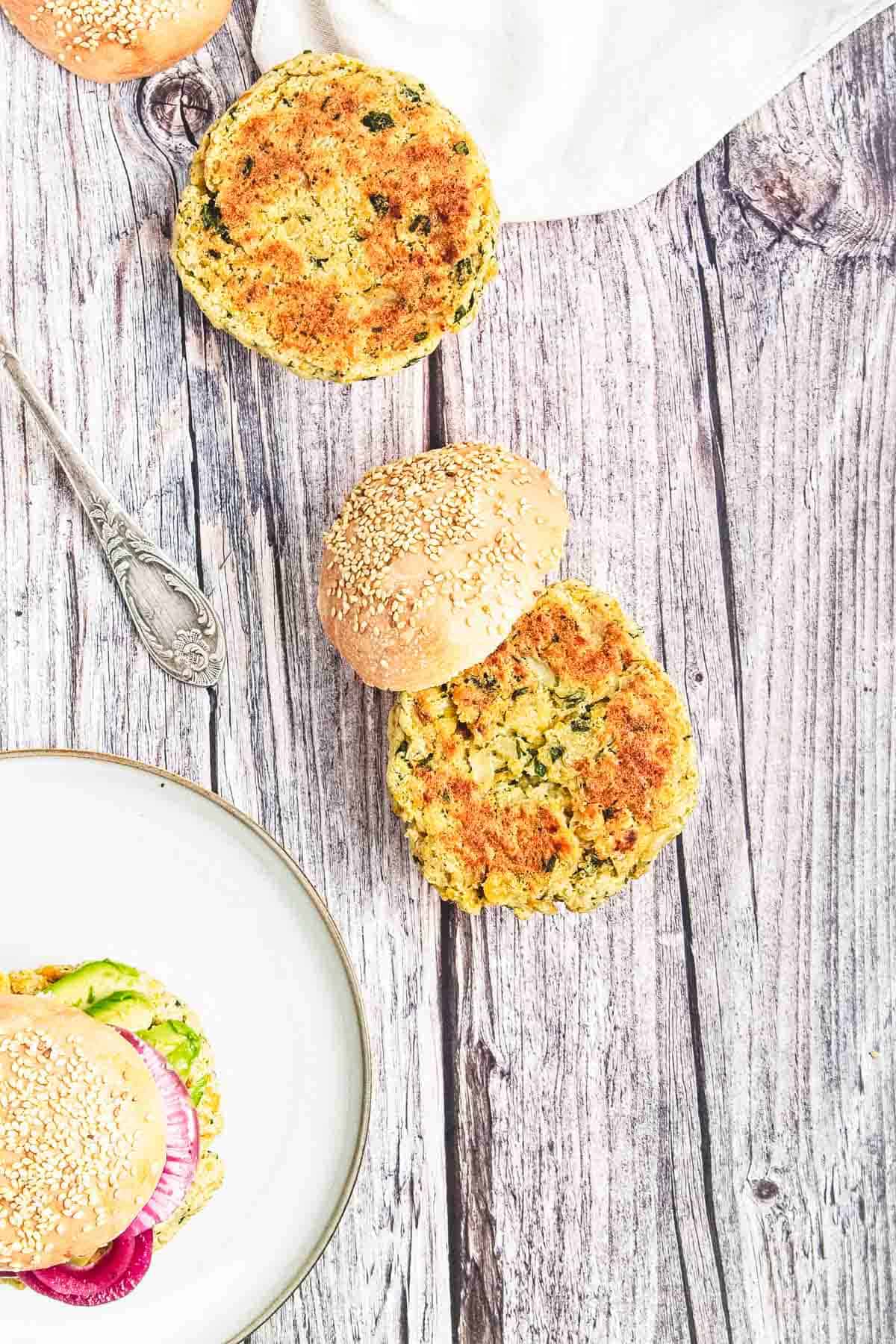 Two chickpea burgers on a sesame bun rest on a rustic wooden surface; another burger is partially visible on a plate.