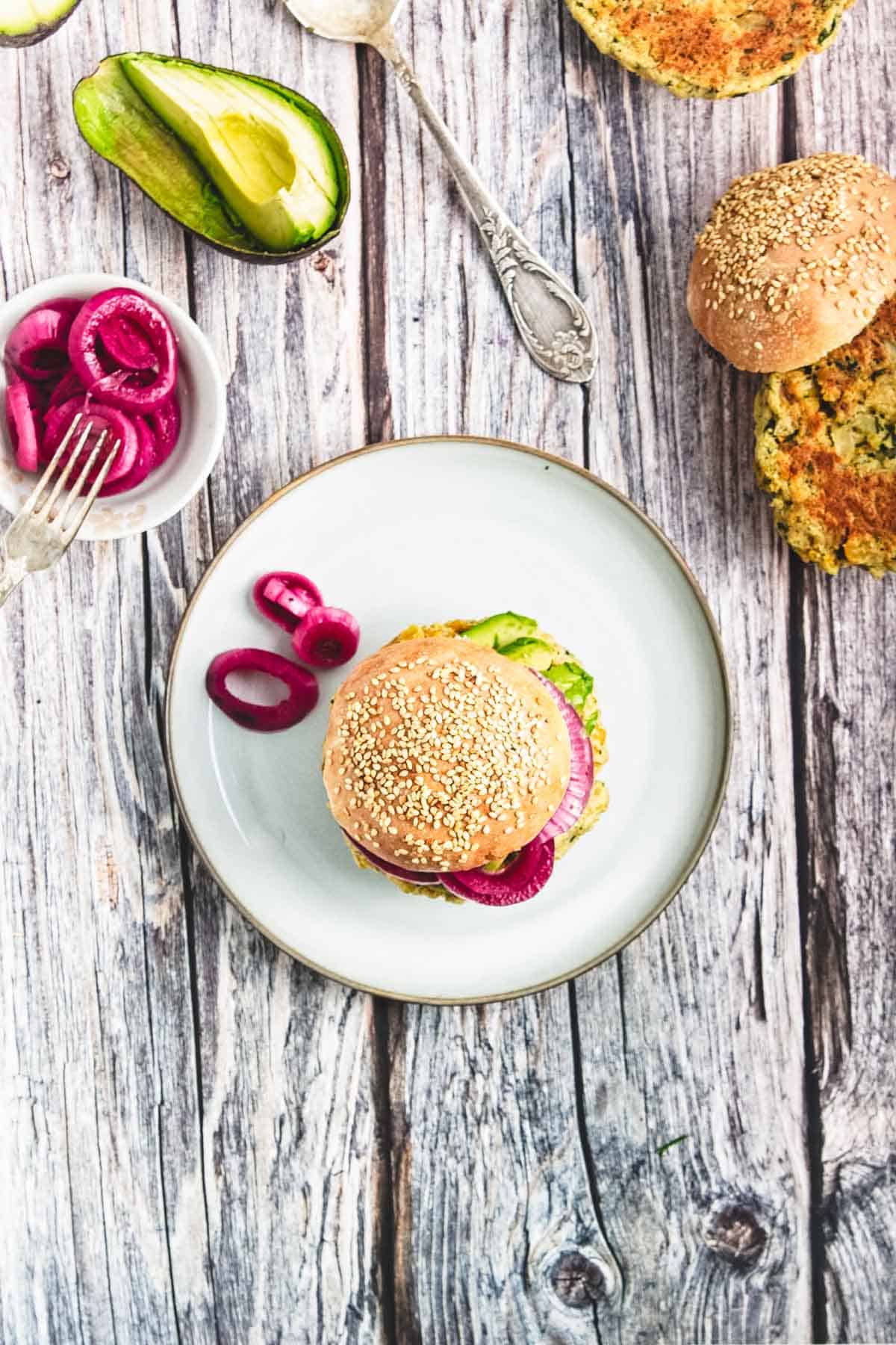 A chickpea burger with avocado and pickled onions sits on a plate, surrounded by extra toppings and a bun on a rustic wooden table.