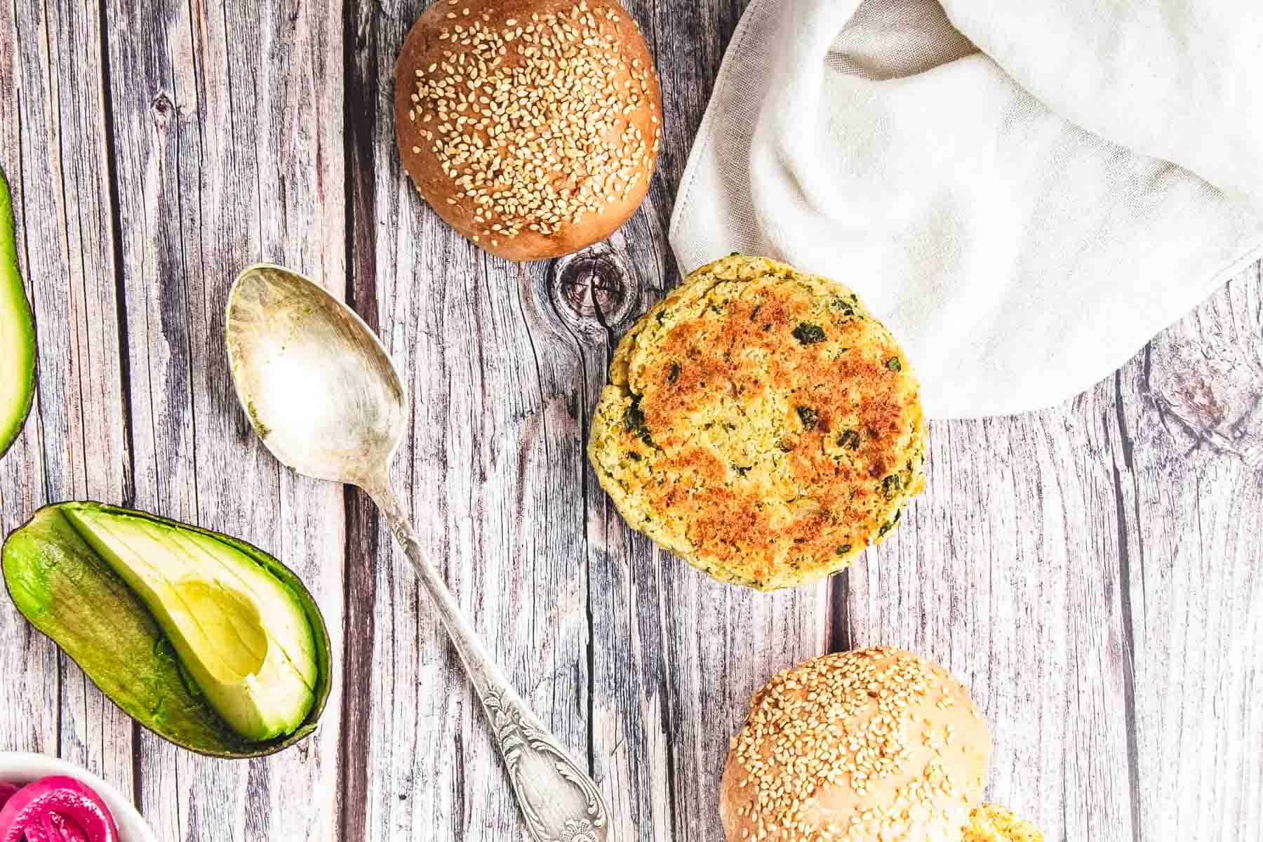 Overhead view of bread rolls, a veggie patty—resembling chickpea burgers—avocado, and a spoon on a rustic wooden surface.