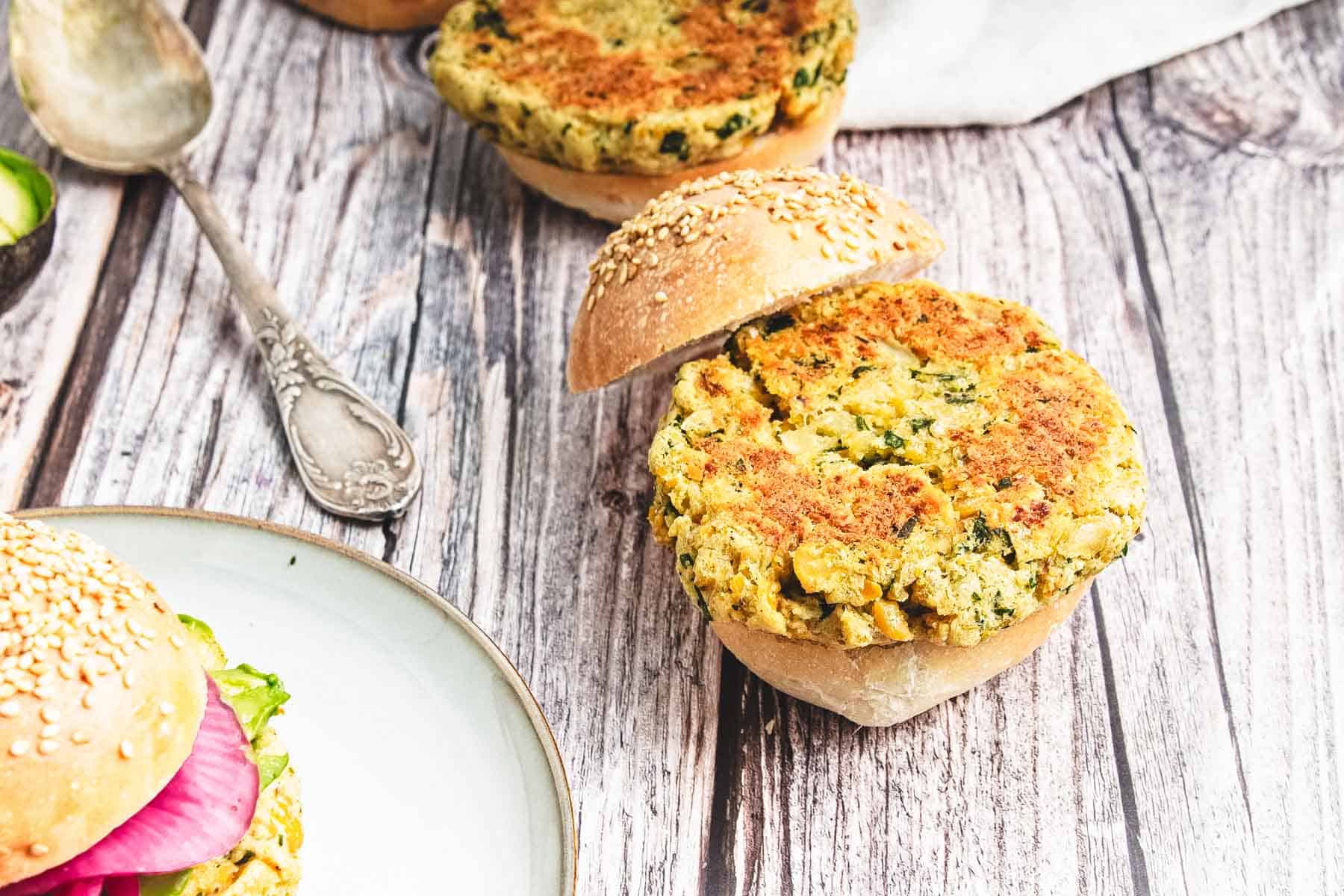 A chickpea veggie patty in a sesame bun sits open-faced on a rustic wooden table, showcasing the hearty appeal of chickpea burgers, with another sandwich nearby.