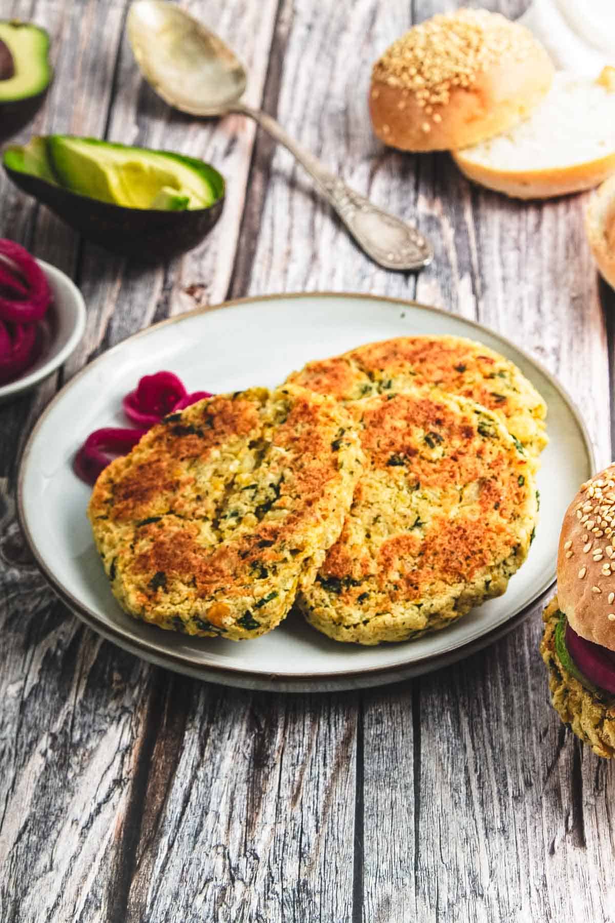 Two chickpea burgers sit on a plate with pickled onions, surrounded by fresh buns and creamy avocado on a rustic table.