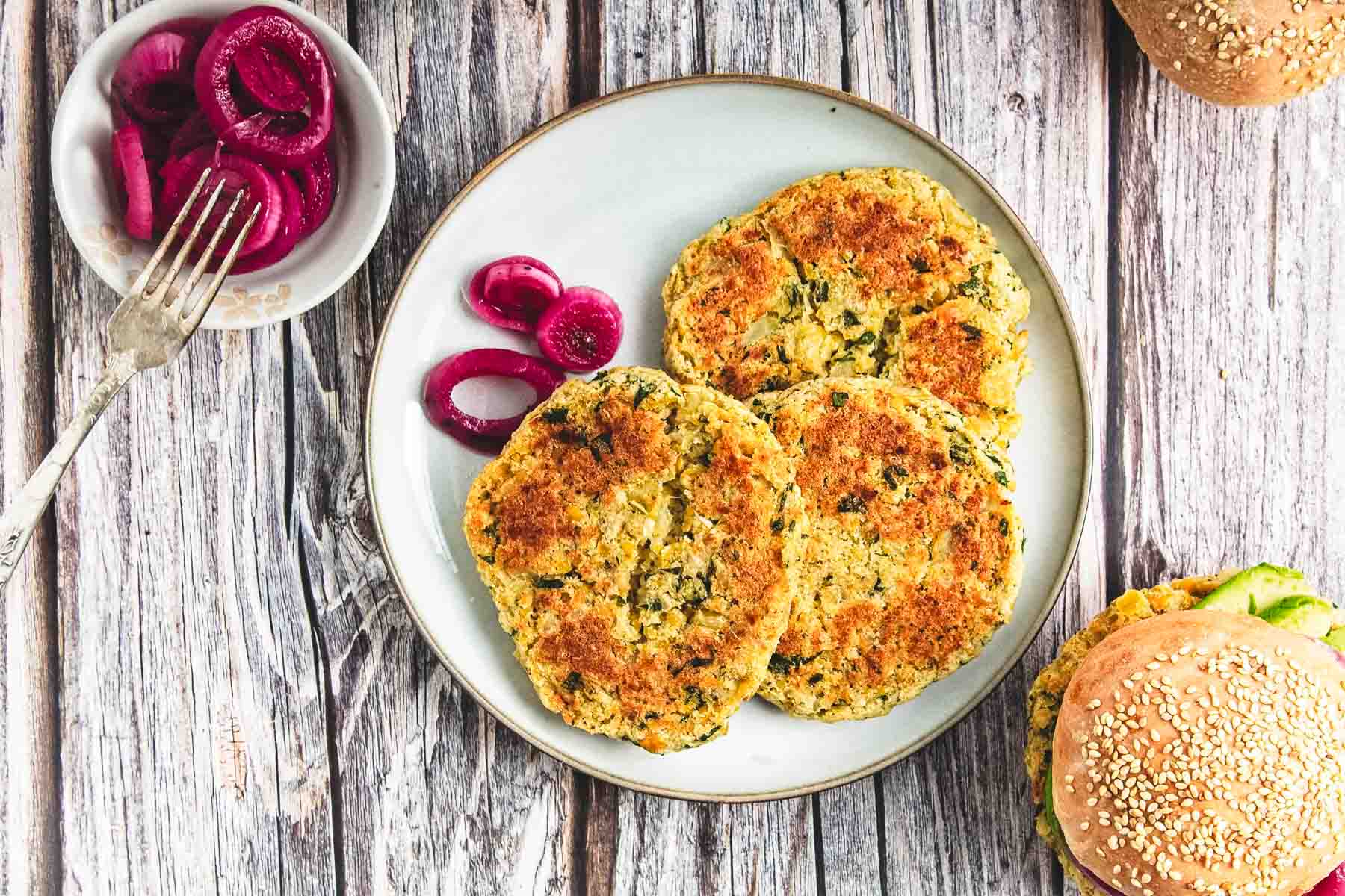 Three golden-brown chickpea burgers on a plate with pickled onions, alongside a classic burger and extra onions on the side.