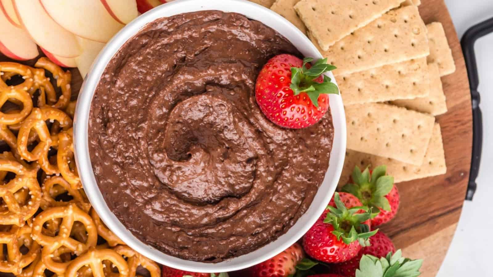 A bowl of chocolate dip surrounded by strawberries, pretzels, apple slices, and graham crackers.