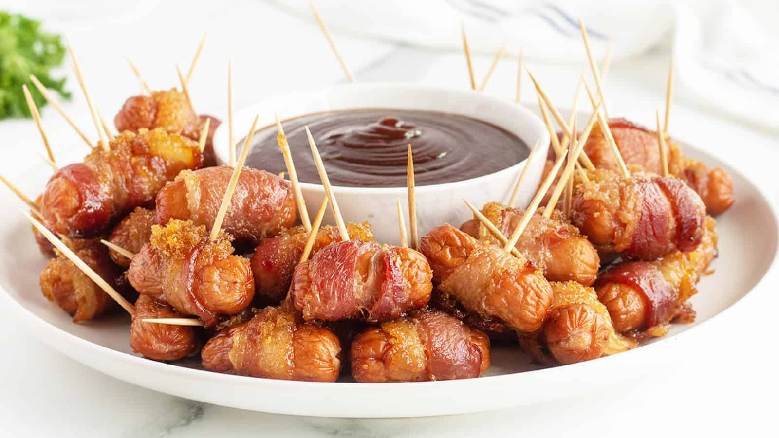 Mini sausages wrapped in bacon with toothpicks, served with a bowl of barbecue dipping sauce on a white plate.