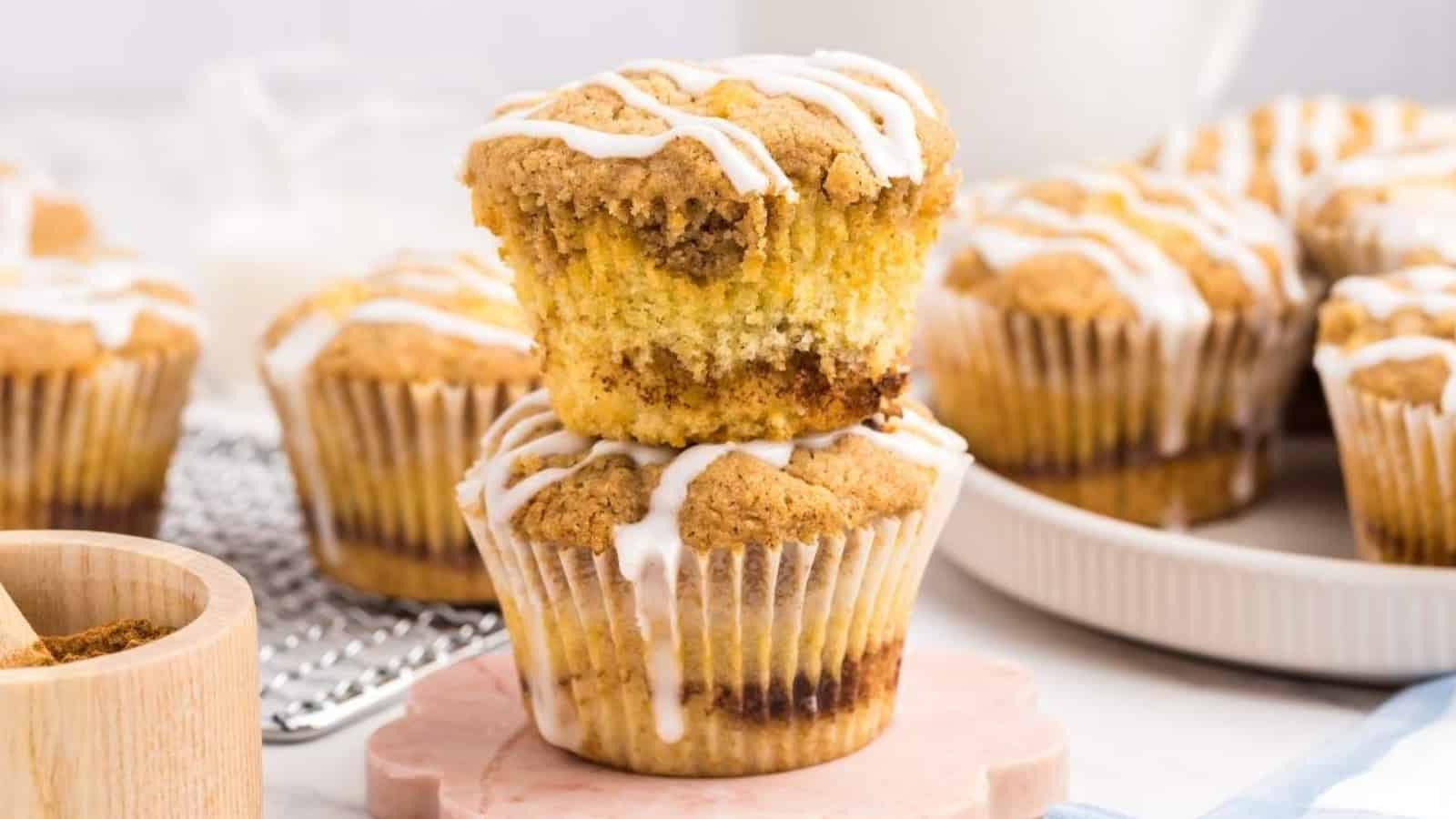 Two cinnamon crumb muffins stacked, drizzled with white icing, with more muffins in the background.