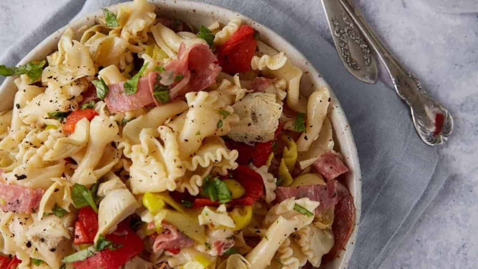 Pasta salad with salami, peppers, and basil in a white bowl, served on a gray napkin with silverware.