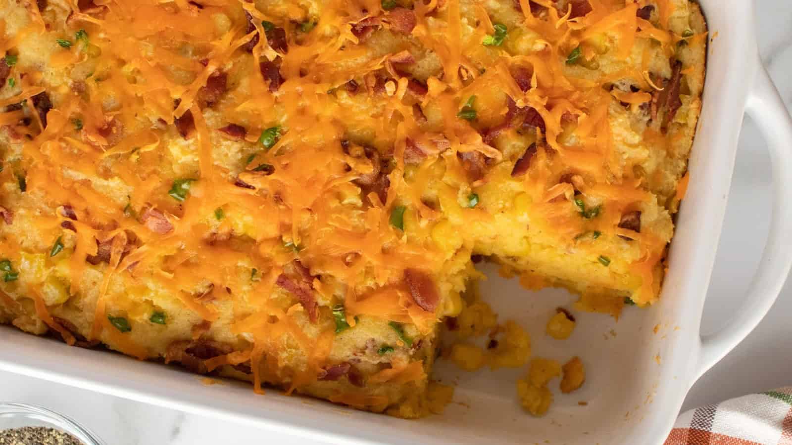 Baked casserole with melted cheese, corn, and green peppers in a white dish with a serving removed.