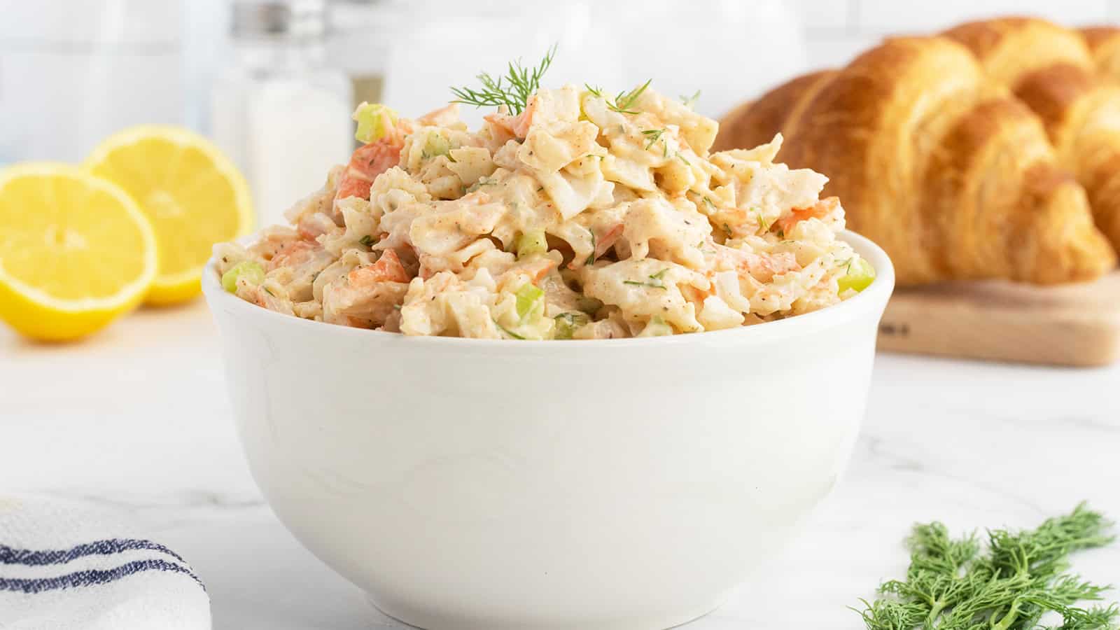 White bowl filled with creamy seafood salad, garnished with dill, with croissants and lemons in the background.