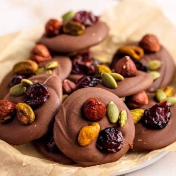 Chocolate disks topped with dried cranberries, pistachios, hazelnuts, and pumpkin seeds on parchment paper.