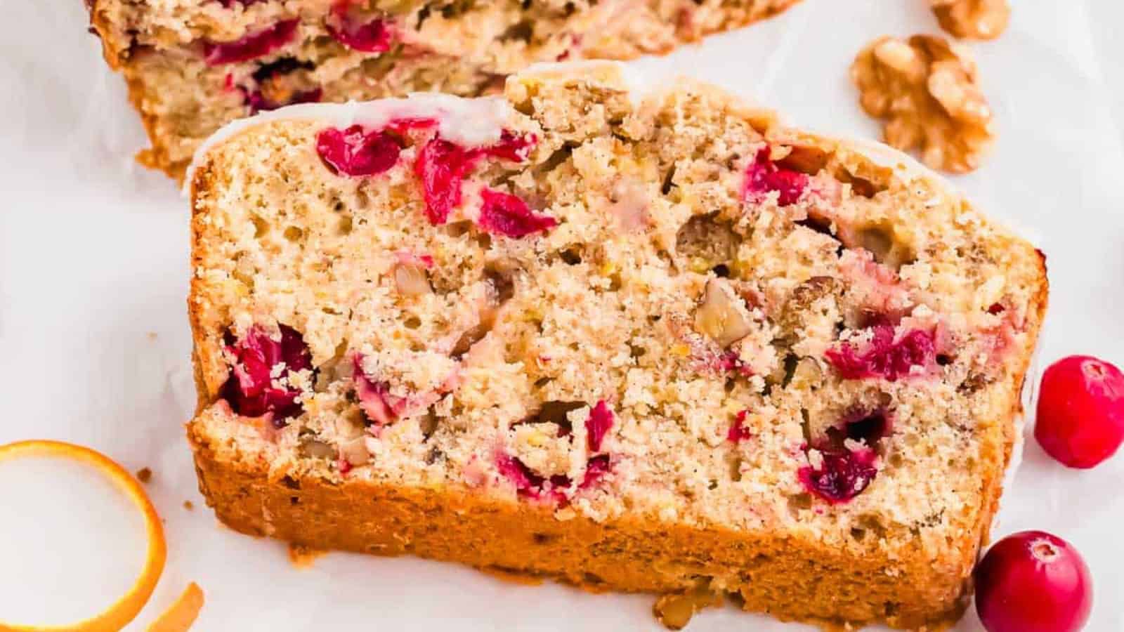 Slice of cranberry nut bread with visible cranberries and walnuts, set on a white surface with whole cranberries nearby.