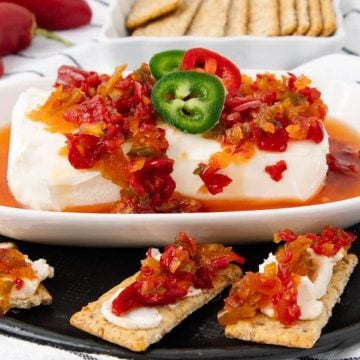 Block of cream cheese topped with pepper jelly and sliced jalapeños, served with crackers on a black plate.
