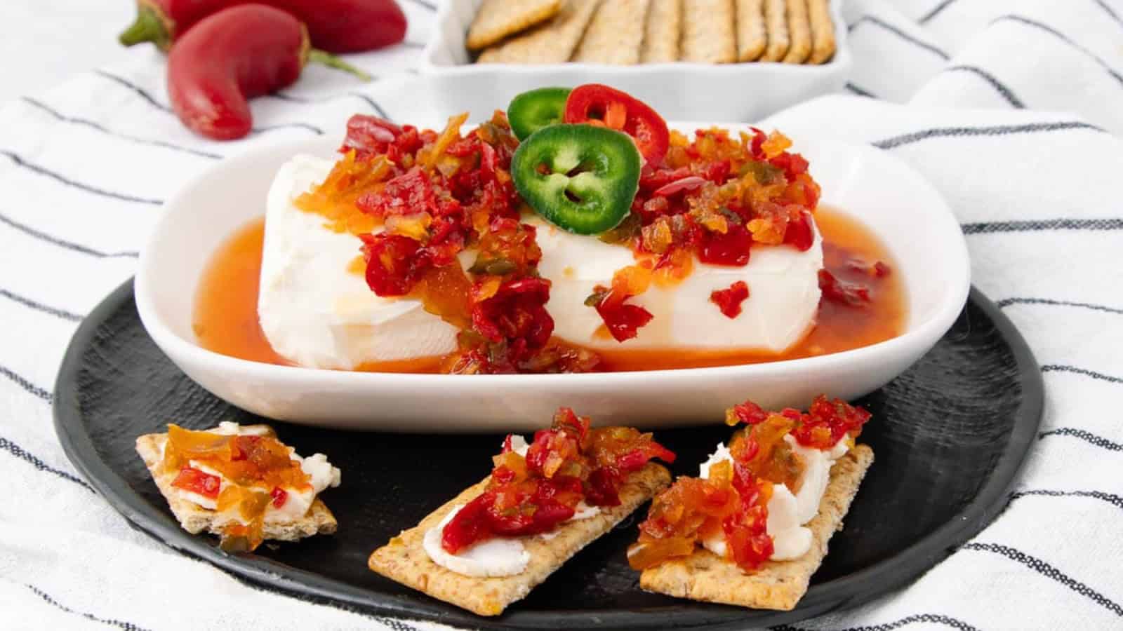 Block of cream cheese topped with pepper jelly and sliced jalape&ntilde;os, served with crackers on a black plate.