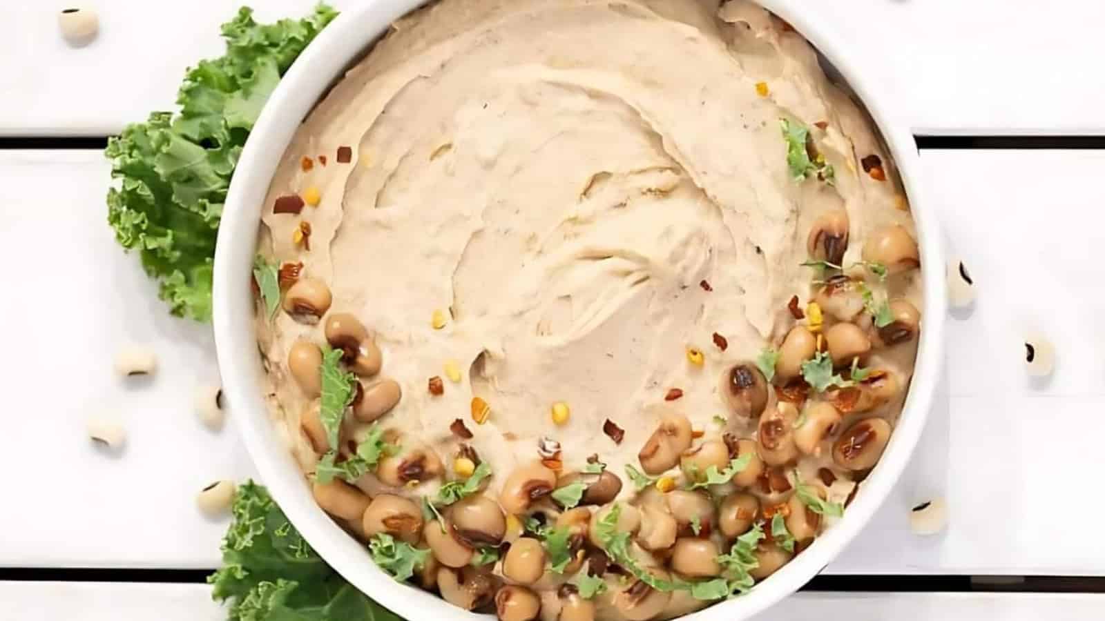 A bowl of creamy dip topped with black-eyed peas, herbs, and red pepper flakes, garnished with leafy greens.