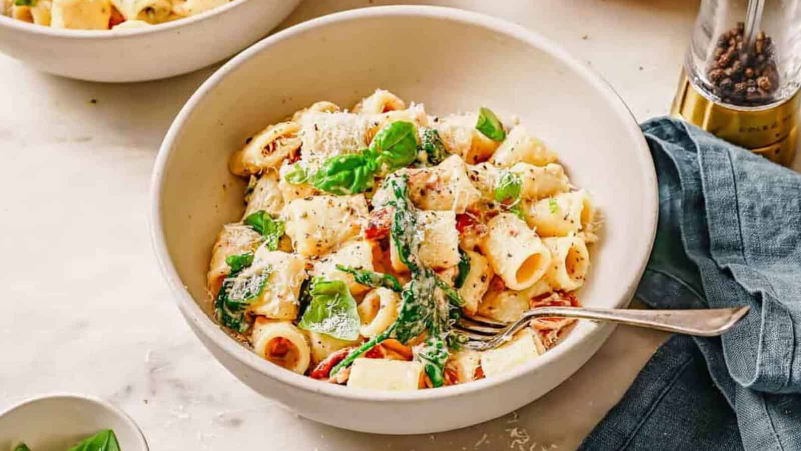 A bowl of rigatoni pasta with spinach, sun-dried tomatoes, parmesan, and a fork on the side.