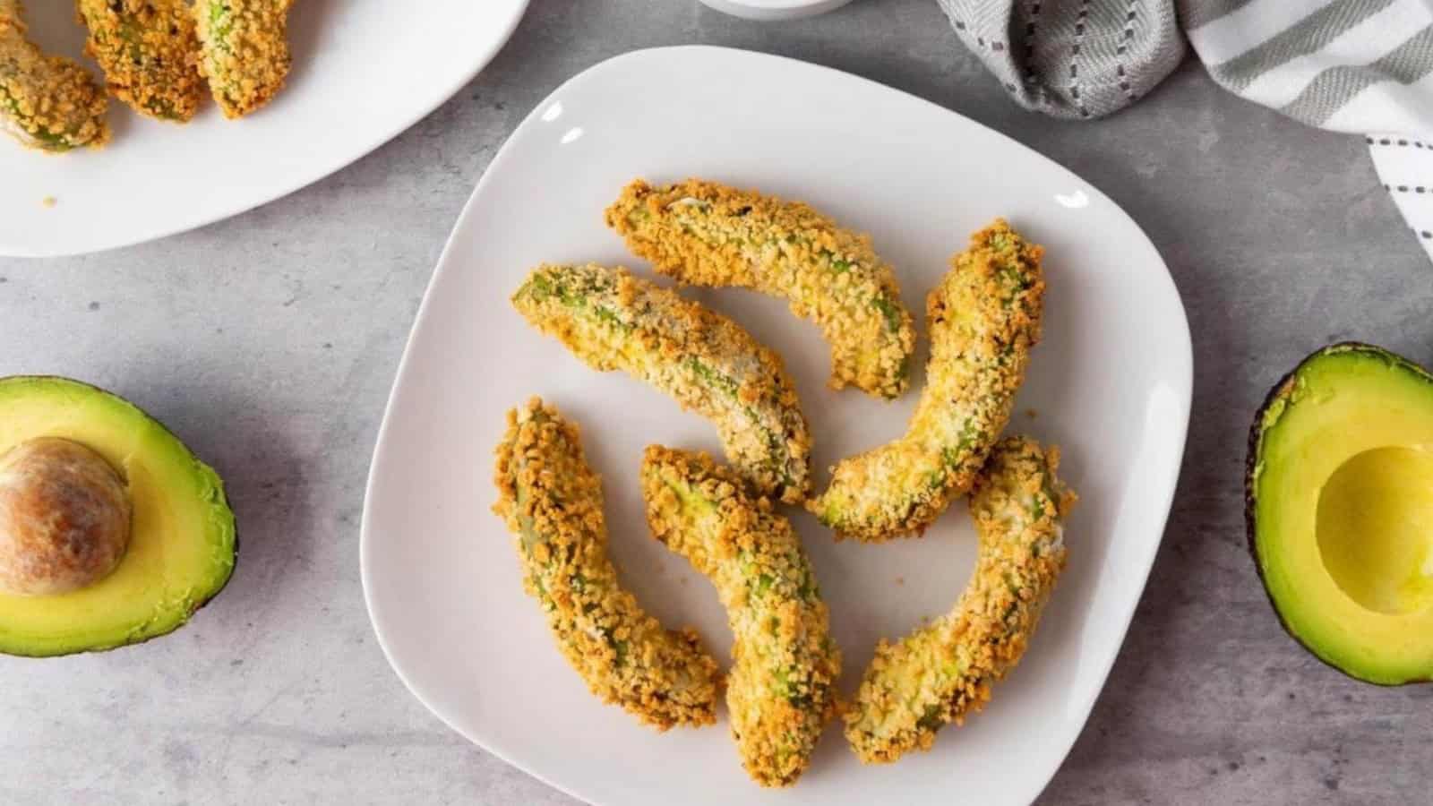 Six breaded avocado slices are arranged on a white plate, with fresh avocado halves nearby.