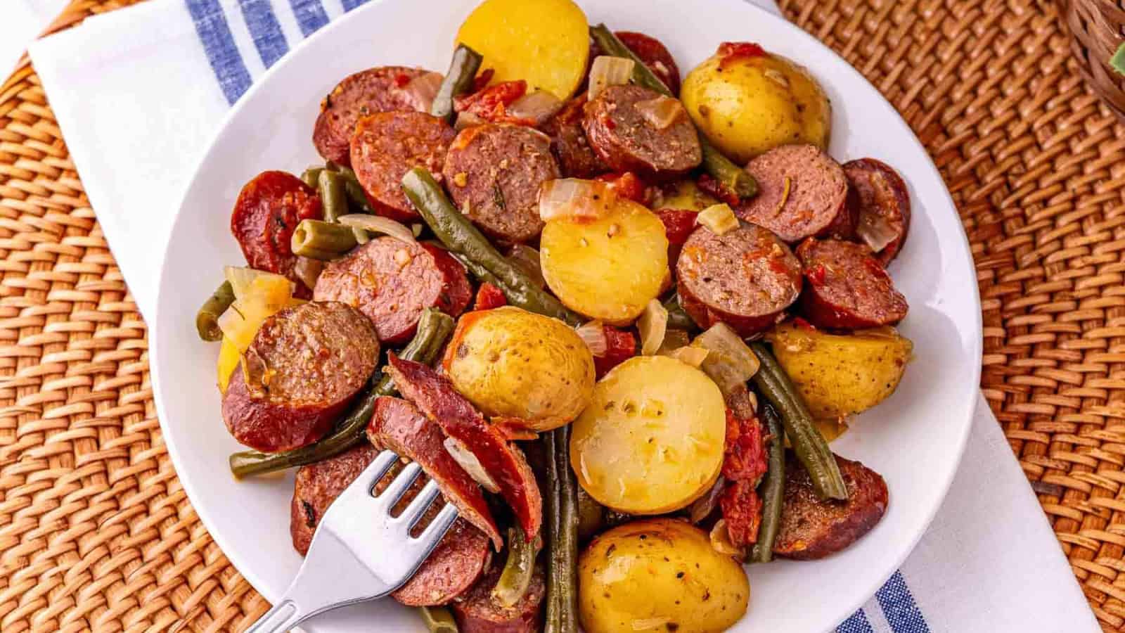 A plate of sliced sausage, potatoes, green beans, and onions, with a fork resting on the side.