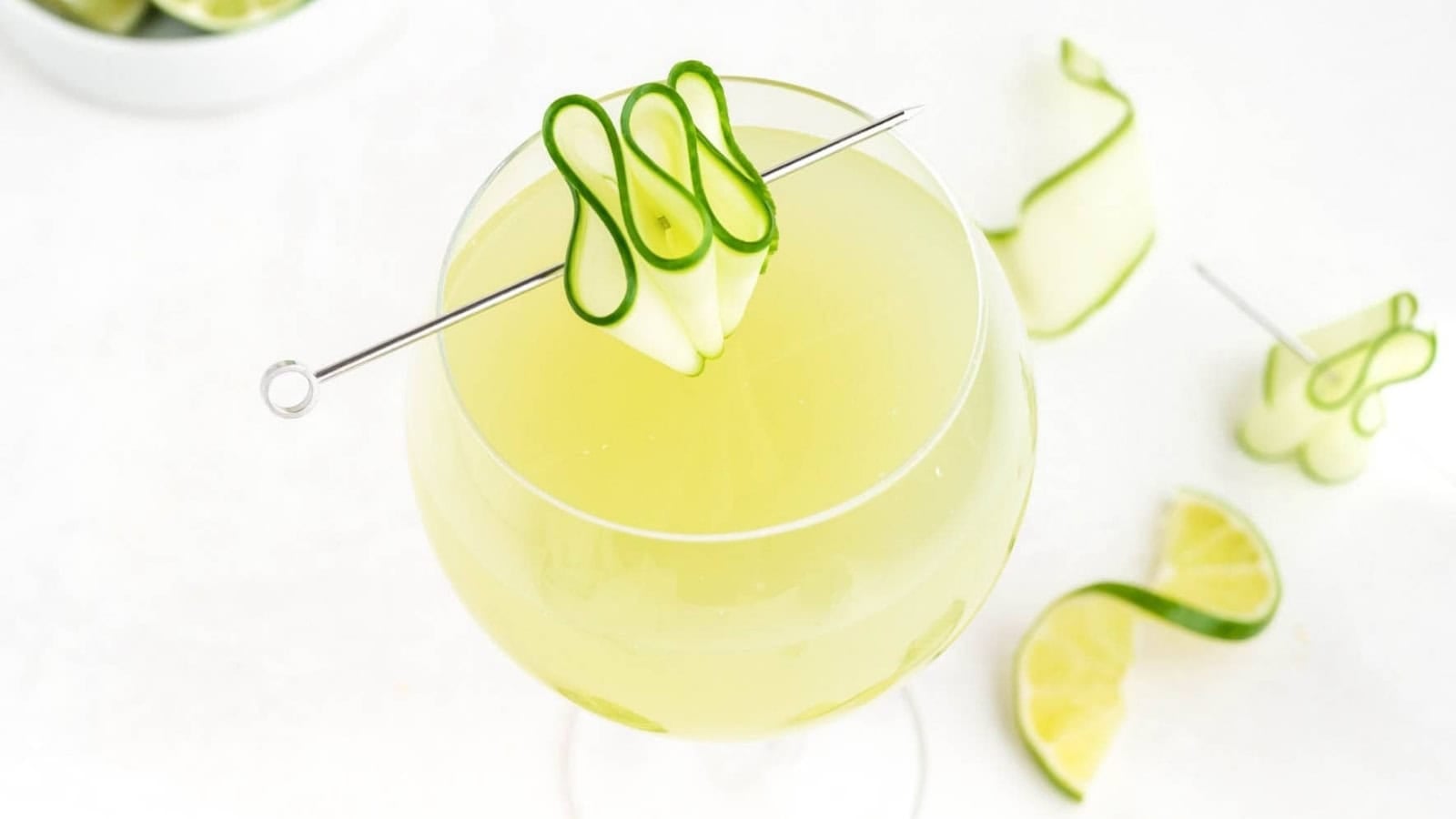 A yellow-green cocktail in a glass, garnished with a curled cucumber ribbon on a metal pick.