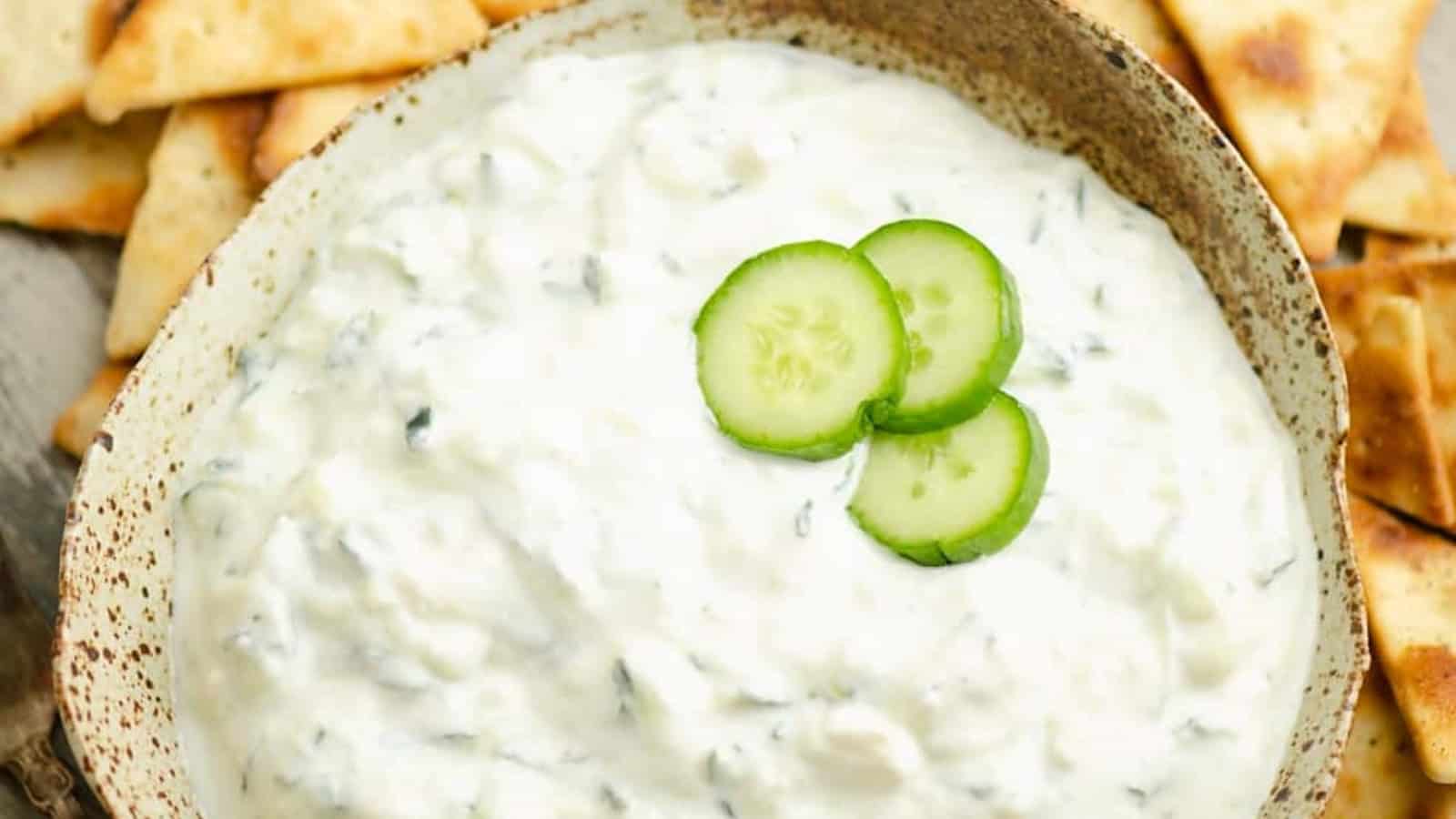 Bowl of tzatziki dip topped with cucumber slices, surrounded by pieces of pita bread.