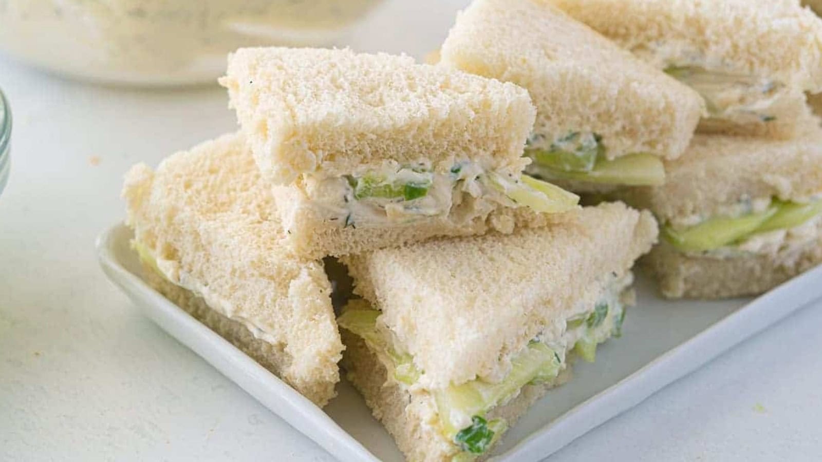 A plate of triangle-cut cucumber and cream cheese sandwiches with white bread, stacked neatly.