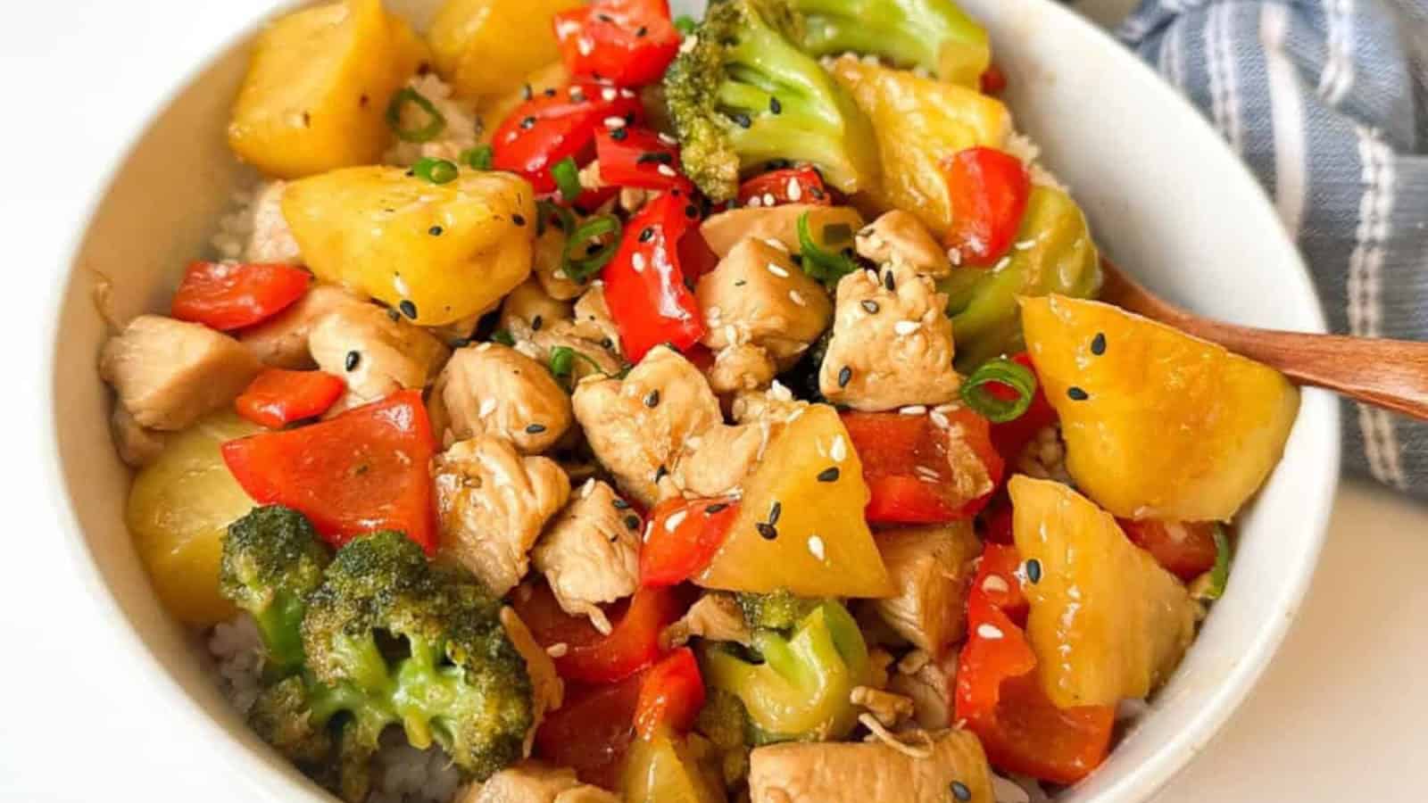 A bowl of chicken stir-fry with broccoli, red bell peppers, pineapple chunks, and sesame seeds on rice.