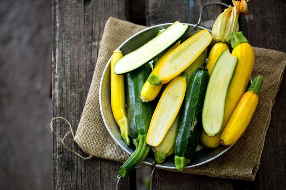 A bowl of sliced yellow and green zucchini on burlap, set on a rustic wooden table.