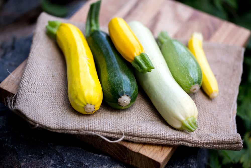 Assorted yellow, green, and pale zucchini on burlap atop a wooden board outdoors.