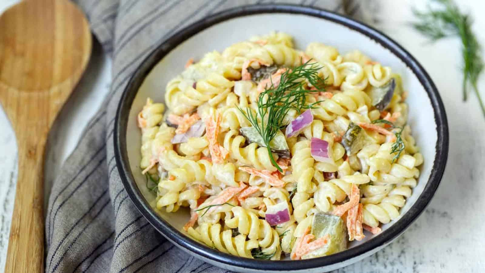 A bowl of creamy pasta salad with rotini, carrots, pickles, red onions, and dill garnish.