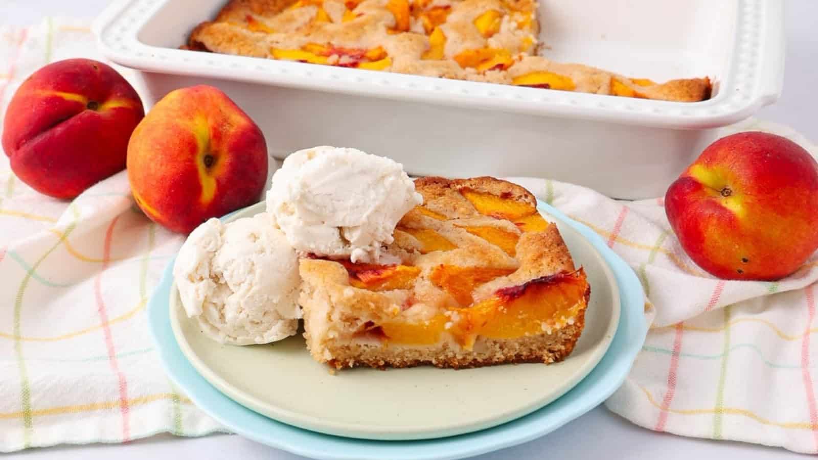 A slice of peach cobbler with two scoops of vanilla ice cream, fresh peaches, and a baking dish in the background.