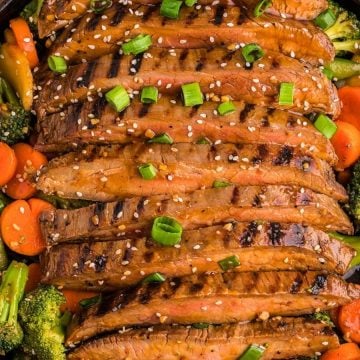 Sliced grilled steak topped with sesame seeds and green onions, served with broccoli and carrots in a skillet.
