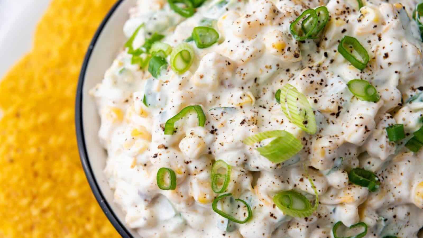 Creamy corn salad topped with sliced green onions and cracked black pepper, next to a pile of corn chips.